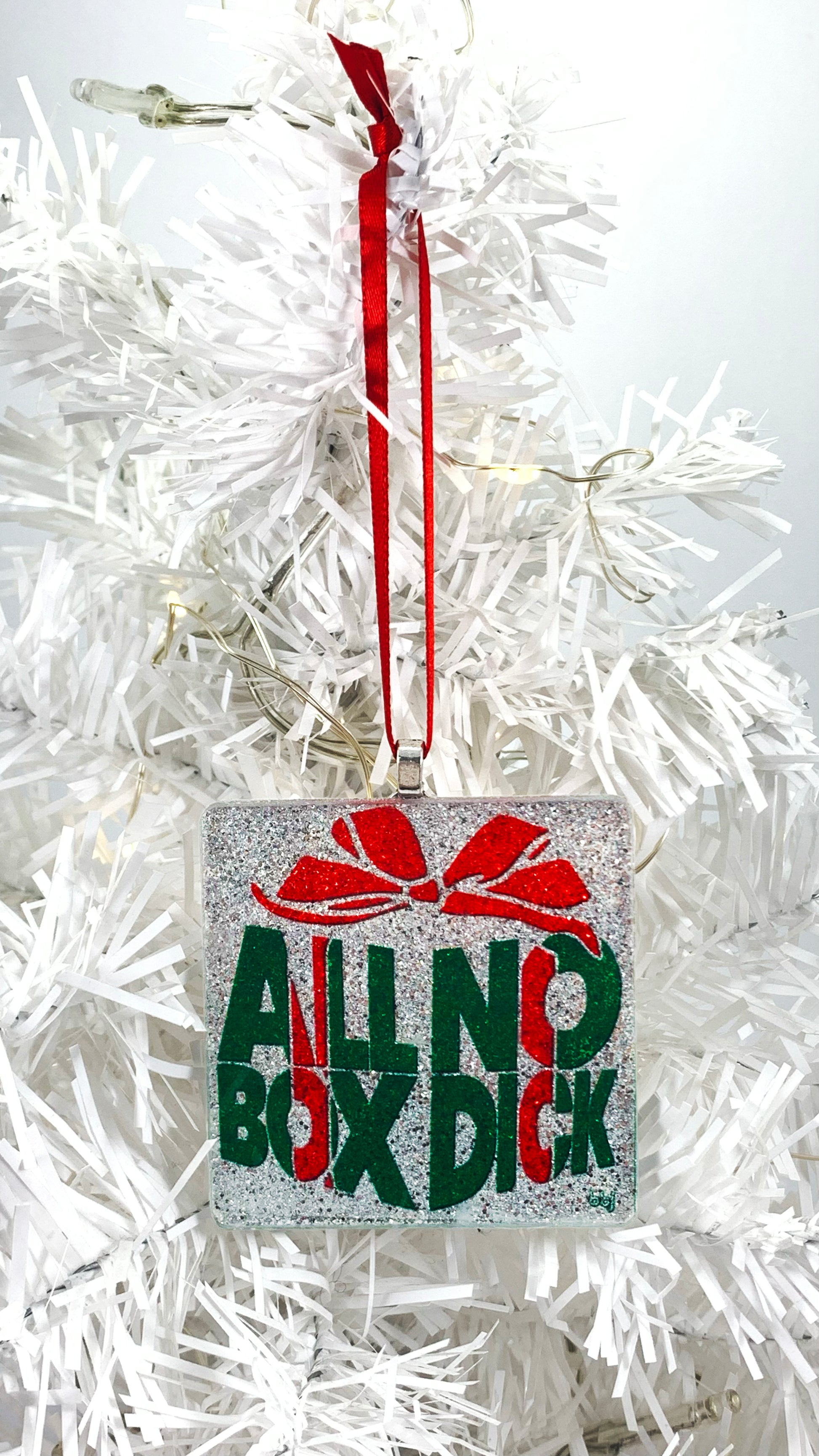 handmade glass ornament with "All Box No Dick" text-based gift-box-shaped graphic design in red and green on silver underlay - tall view with complete red ribbon, hanging on white Christmas tree