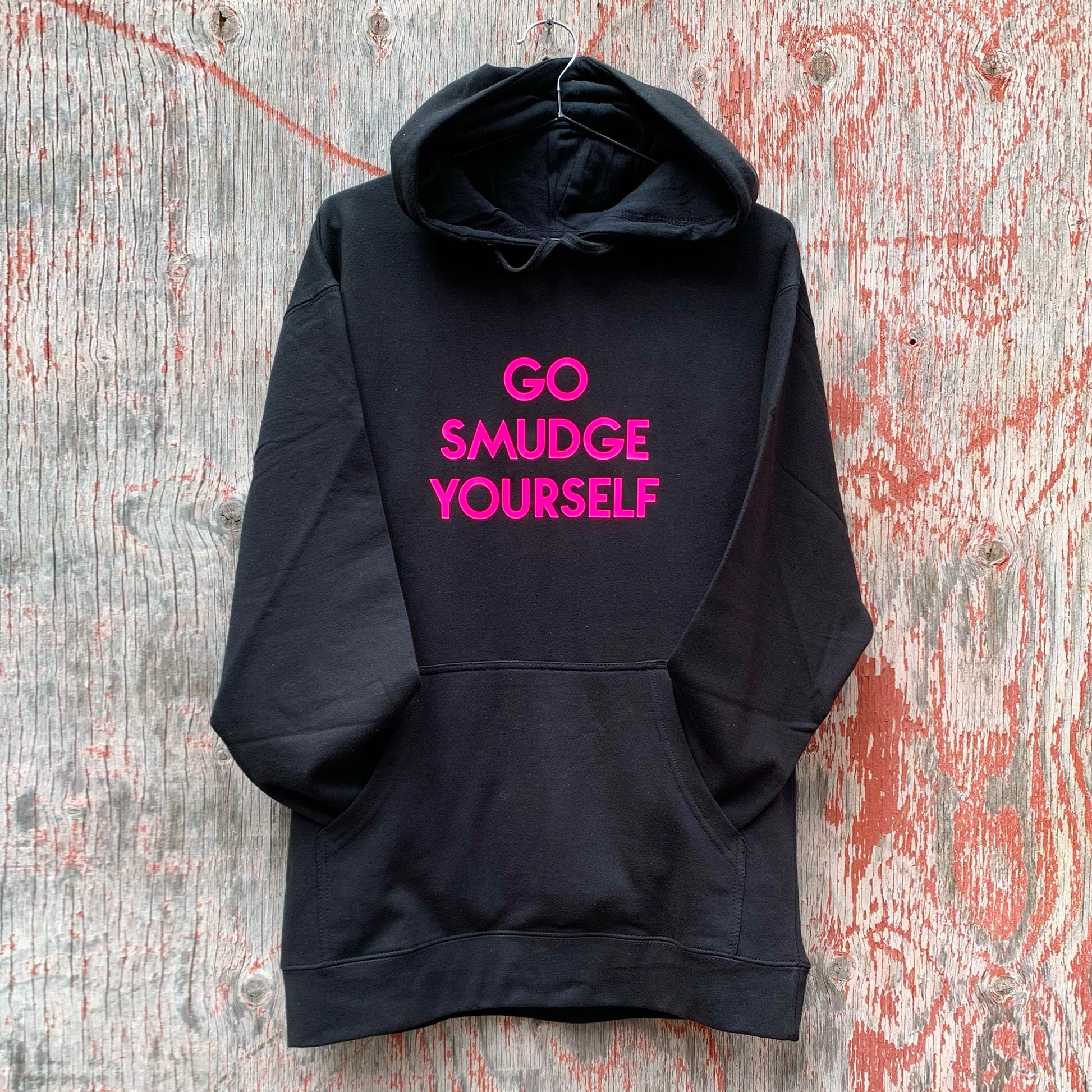 photo of custom black hooded sweatshirt with "Go Smudge Yourself" in neon pink geometric text, hanging on weathered wood